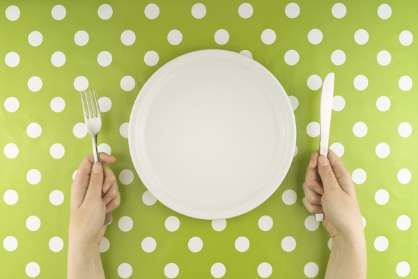 Female hands at dinner table holding fork and a knife above empty flat white plate, dieting concept.
