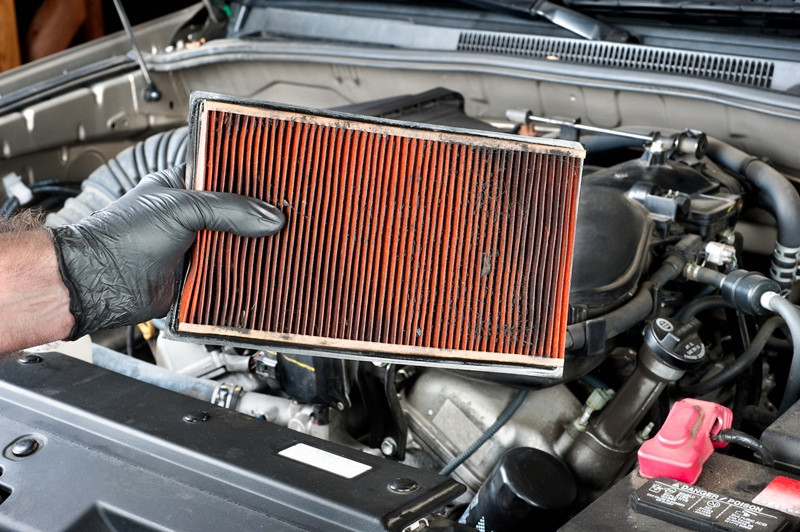 An auto mechanic wearing protective work gloves holds a dirty, clogged air filter over a car engine during general auto maintenance.